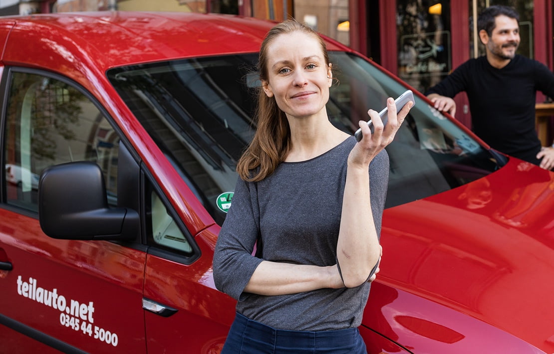Zwei Personen stehen an einem roten teilAuto-Fahrzeug, eine hat ein Smartphone in der Hand